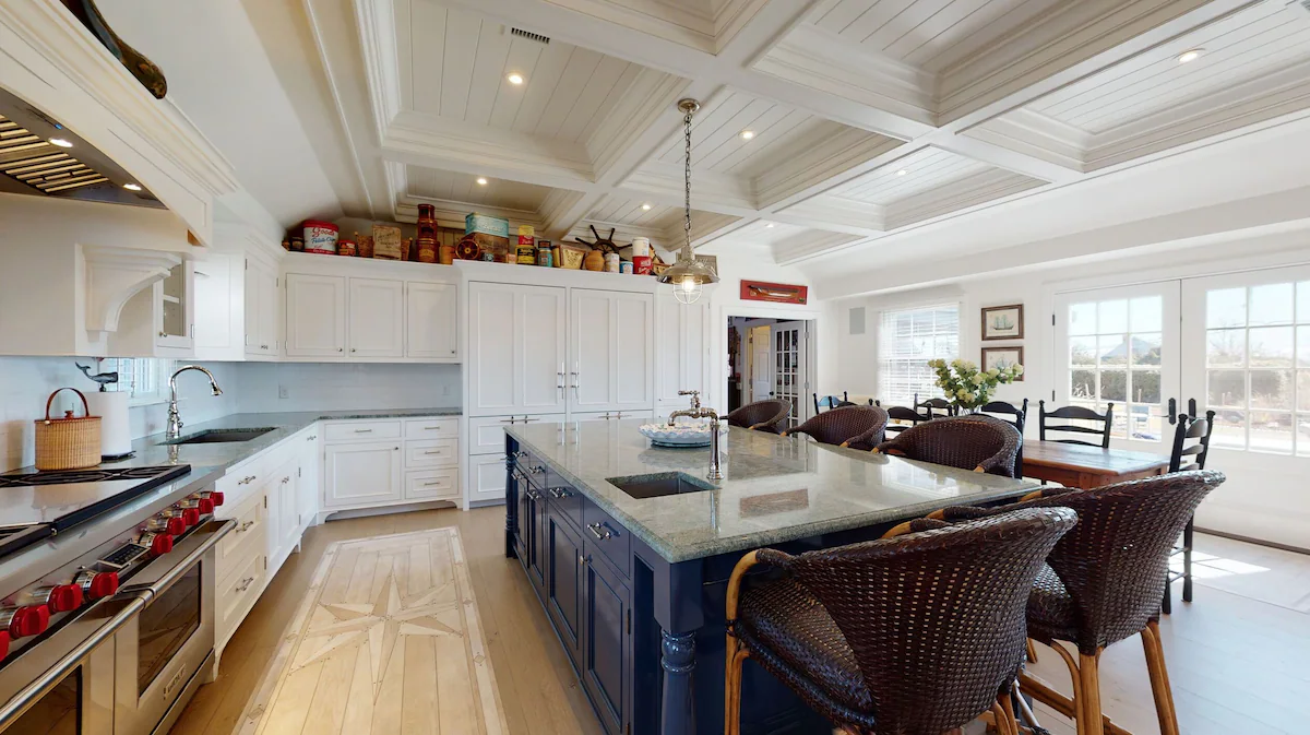 The range wall from the far side of the island: a professional range under a beam-and-corbel hood, floor-to-ceiling paneled cabinetry, antique tins and crocks lined along the cabinet tops, and a compass rose inlaid into the pale wood floor