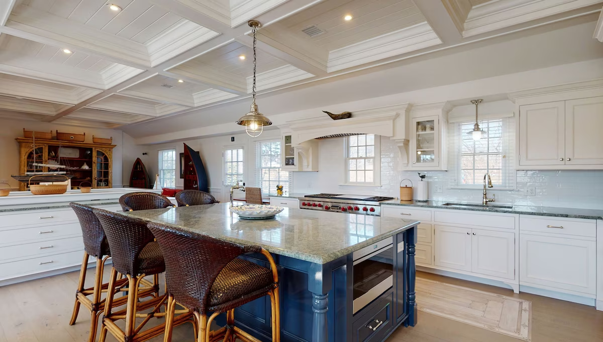 The kitchen looking across a long navy island with green granite counters and six woven barstools, toward a professional range with red control knobs under a shingled hood, white cabinetry, and a coffered white plank ceiling