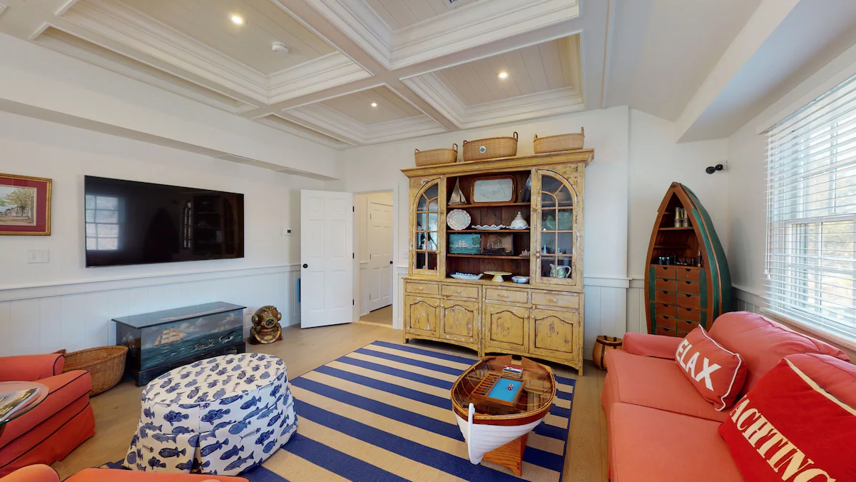 The other end of the living room: a wall-mounted television, a distressed yellow glass-front hutch topped with Nantucket baskets, a hand-painted ship chest, and a second dory fitted with drawers beside the window