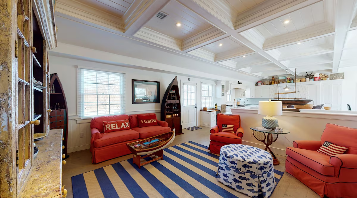 The living room: coral slipcovered sofa and armchairs with RELAX and YACHTING pillows on a navy-and-cream striped rug, a wooden rowboat coffee table, a fish-print ottoman, and a dory hull fitted out as a bar, open to the kitchen beyond