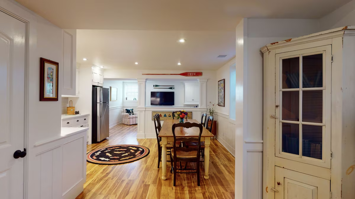 The same rooms from the far doorway, with a distressed cream corner cupboard in the foreground, a hooked oval rug under the farm table, and maple floors running through to the sofa at the end