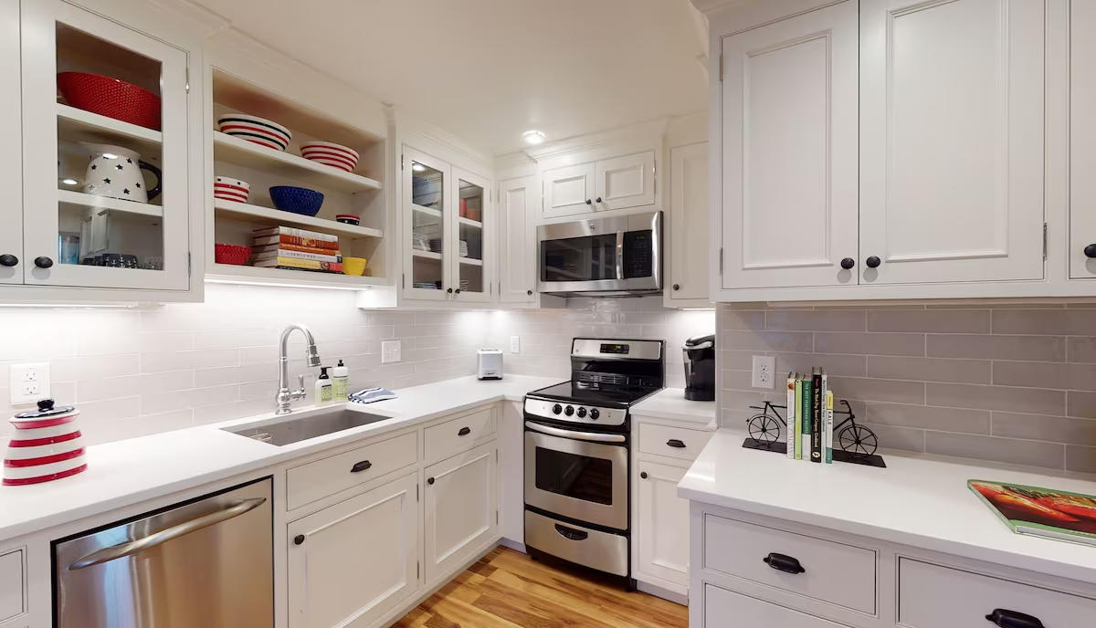 The lower-level kitchen corner: glass-front and open shelving stacked with red and navy dishware, gray subway tile, white quartz counters, a stainless range and a dishwasher
