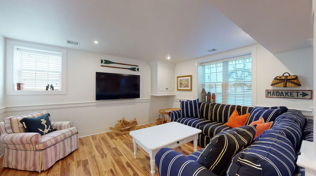 The lower-level family room: a navy-and-white striped sectional with orange pillows, a striped armchair with an anchor pillow, a wall-mounted television under crossed green oars, a MADAKET arrow sign and a wicker creel on the wall