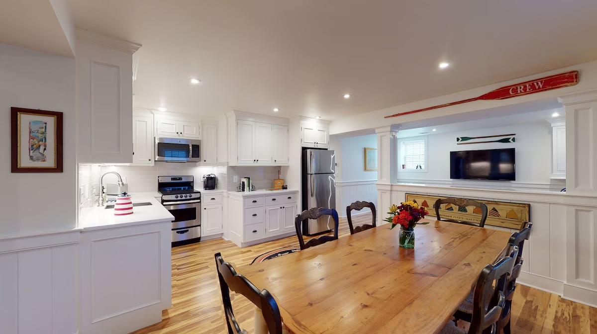 The lower-level kitchen open to the dining table: white shaker cabinetry, a stainless range and refrigerator, a reclaimed plank table with antique chairs and a jar of flowers, and a red CREW oar mounted over the pass-through to the family room beyond