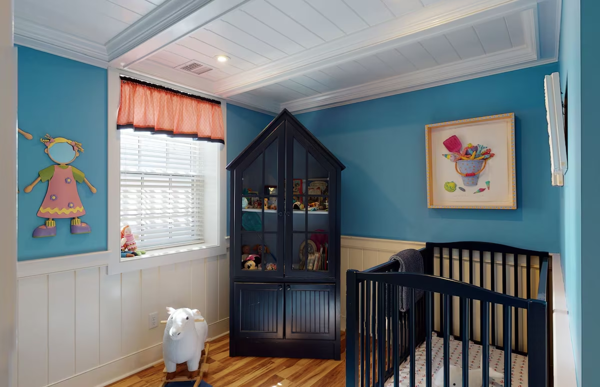 The nursery: teal walls above white beadboard wainscot under a white coffered plank ceiling, a navy crib, a tall navy glass-front armoire filled with toys and books, a wooden paper-doll figure on the wall, a coral valance over the shuttered window, and a white lamb rocker on the wood floor