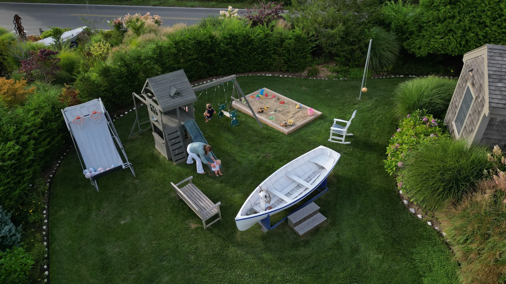 Another aerial angle of the play yard showing a stand-up ball-toss game, the slide-and-swing fort, the sandbox, the rowboat on its cradle with wooden steps, and a teak bench on the lawn