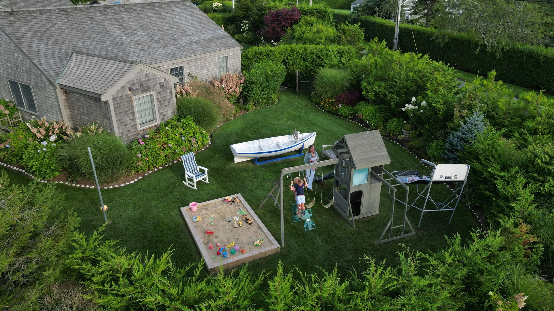 Aerial view of the play yard: the white rowboat on its blue cradle, the timber fort with slide and swing set, the room-sized sandbox full of toys, a striped rocking chair and a tetherball pole, framed by deep flower borders and clipped hedges