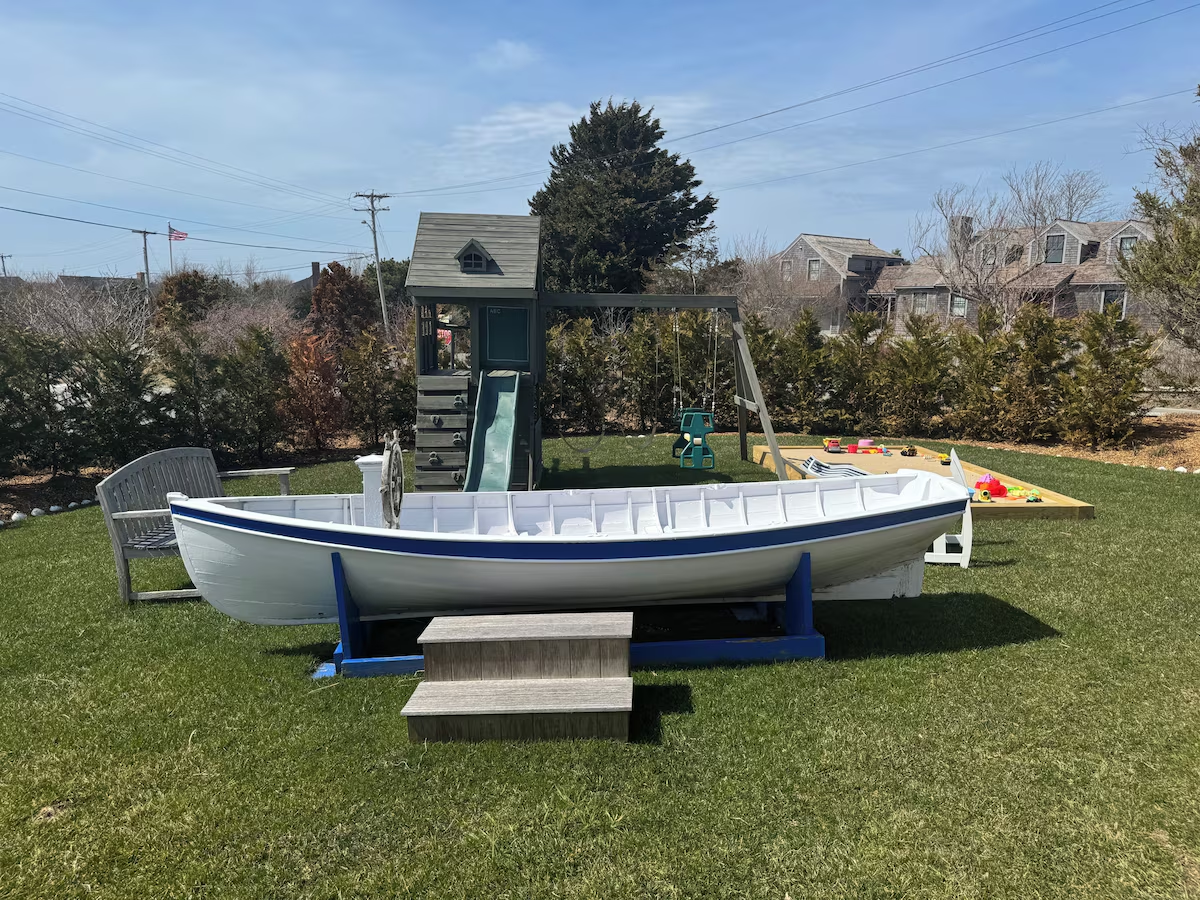The rowboat head-on — a full-size white hull with a navy sheer stripe, open framed interior and a ship’s wheel at the bow — on a blue cradle with weathered wooden steps, the slide-and-swing fort behind it and the sandbox off to the right