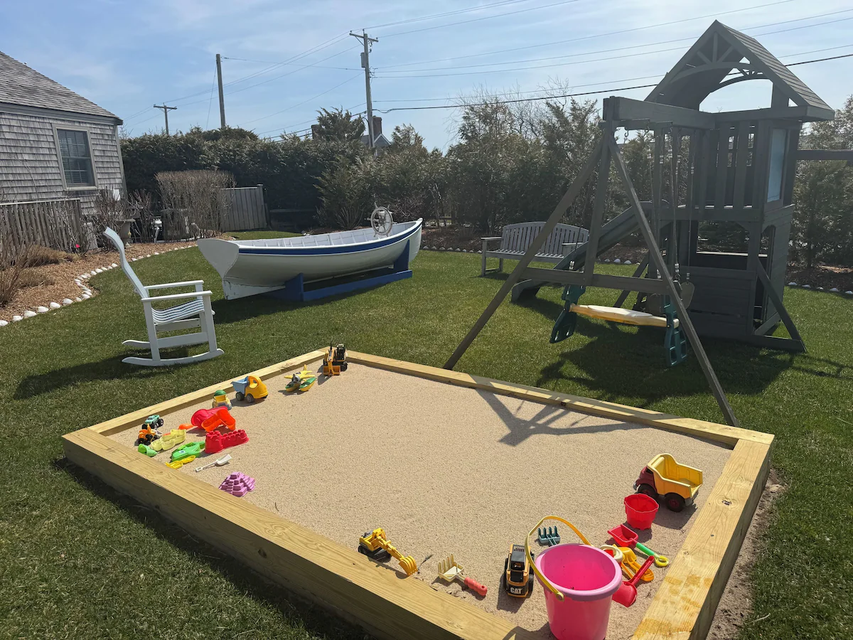 The play corner of the lawn: a large timber-framed sandbox filled with dump trucks, diggers, molds and buckets, a gray wooden fort with a slide and swings beyond it, a white rowboat on a blue cradle, a white rocking chair and a teak bench on the grass