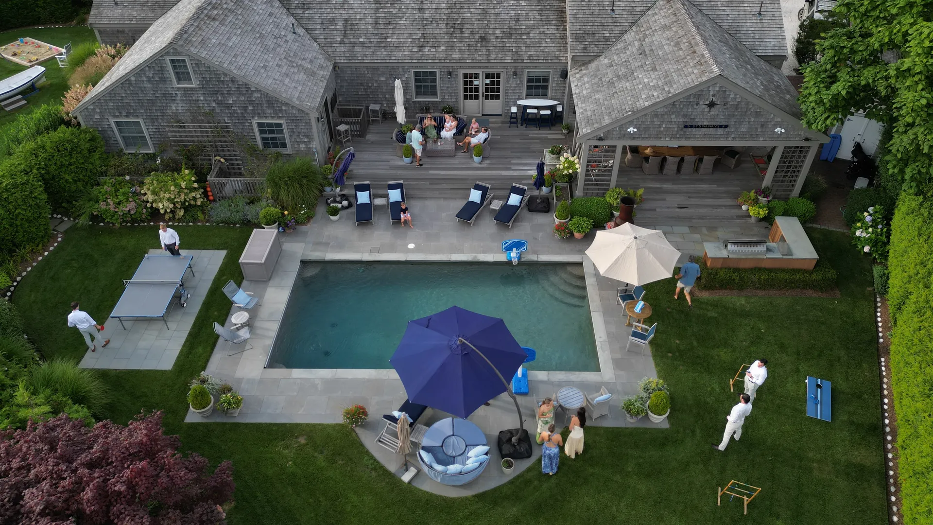 Aerial view of the heated pool on its bluestone terrace: navy chaises and cream and navy umbrellas along the sides, a ping pong table and cornhole boards on the surrounding patio, the built-in grill and the covered porch under the STOWAWAYS nameboard, with guests gathered across the deck