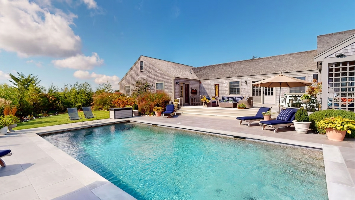 The pool looking back at the house on a clear autumn day: turquoise water inside a pale bluestone surround, navy chaises and a tan umbrella on the right, a wicker sofa with navy striped cushions on the deck above the steps, and cedar-shingled gables behind