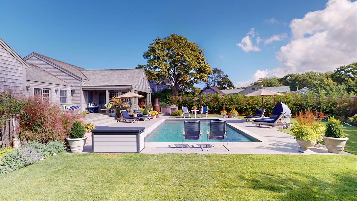 The pool and deck seen across the lawn, with two navy sling chairs at the water’s edge, chaises and umbrellas along both sides, urns of boxwood, and a big shade tree over the shingled ell