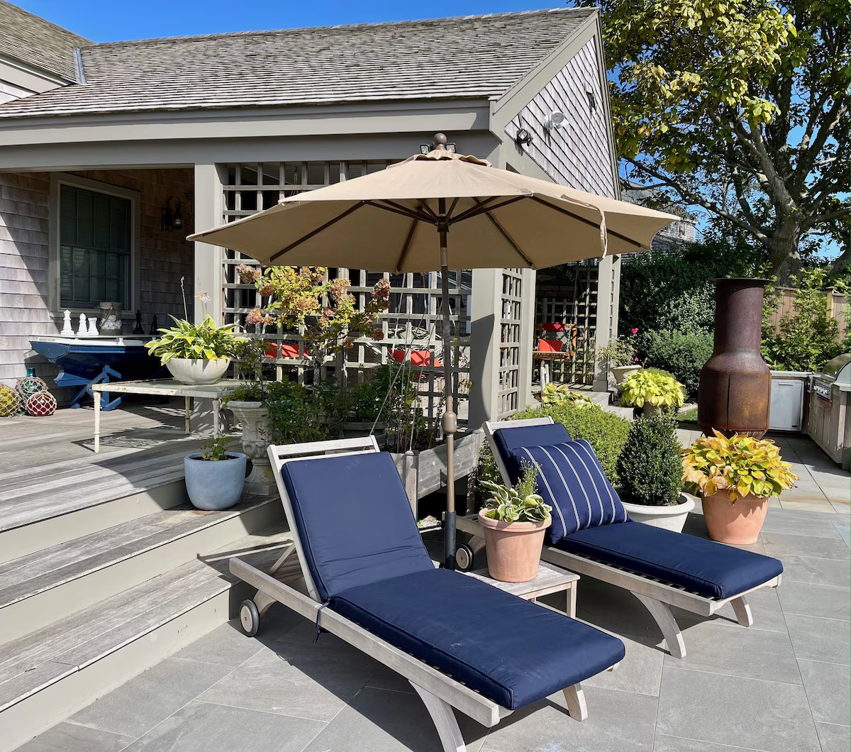 Two teak chaises with navy cushions and a striped pillow under a tan umbrella on the bluestone, a terracotta pot of coleus and clipped boxwood beside them, a rusted steel chiminea and the built-in grill along the deck, and a blue dory table set with chess pieces on the deck above