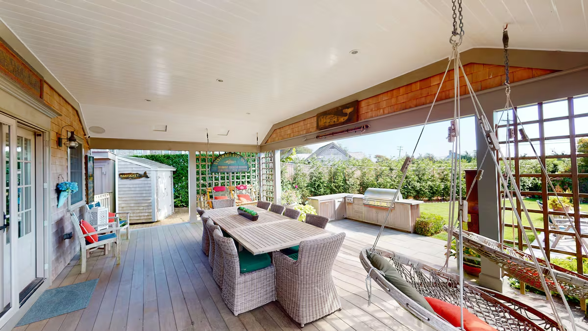 The covered porch: a long wicker dining table for eight with green cushions, a hanging rope swing, a built-in stainless grill, and a white beadboard ceiling over teak decking