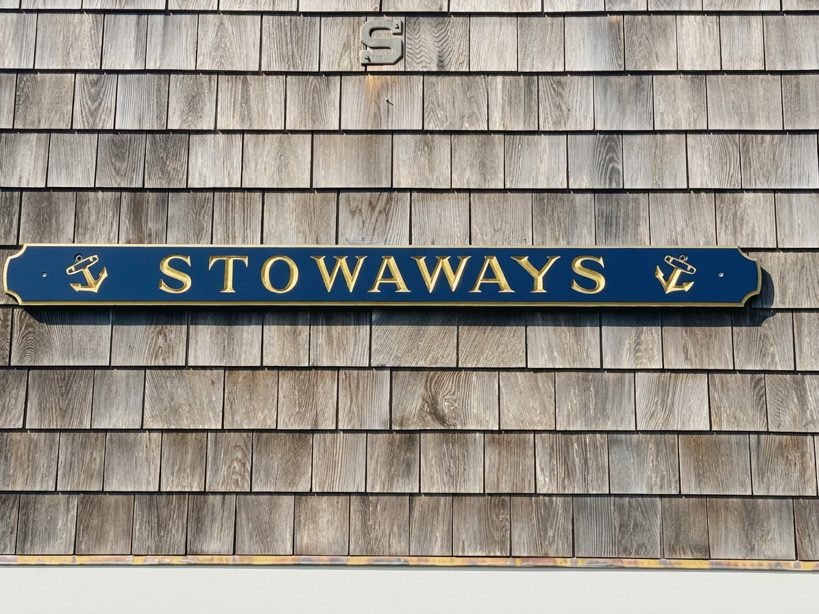 The house’s carved nameboard: "STOWAWAYS" in raised gold serif letters on a navy plank flanked by two gold anchors, mounted on weathered gray cedar shingles.